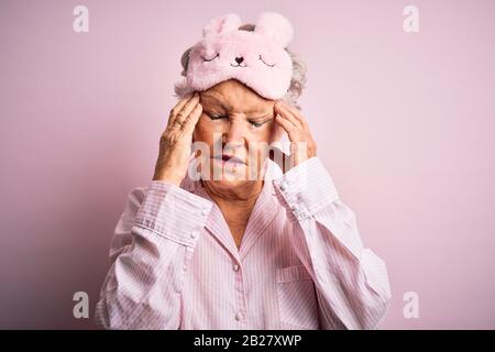 Donna bella anziano che indossa la maschera del sonno e pajama su sfondo rosa isolato con la mano sulla testa per il dolore in testa perché lo stress. La sofferenza migrai Foto Stock