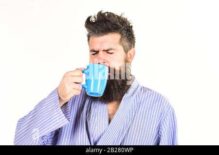 Macho in accappatoio bevande tè o caffè con la faccia del sonno. Ragazzo sembra affogato, ha bisogno di caffè. Caffè del mattino. L'uomo con barba e baffi tiene una tazza di caffè, isolato su bianco Foto Stock