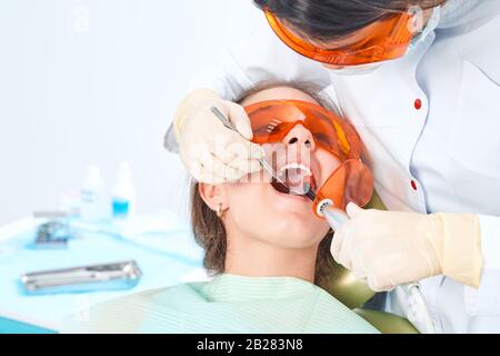 Ragazza bambino al medico. Il dentista posiziona un riempimento su un dente con la lampada di polimerizzazione dentale in cavità orale. Su sfondo clinico Foto Stock