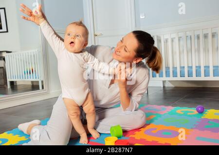 La madre felice gioca con il bambino bambino bambino nella stanza dei bambini aiutandolo a stare in piedi e sollevare le mani Foto Stock