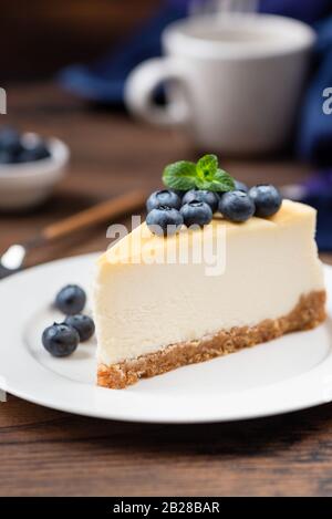 Fetta di cheesecake sormontata da mirtilli su piastra bianca, sfondo tavolo in legno. Composizione verticale. Gustoso pezzo di torta Foto Stock