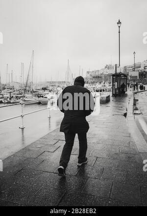 Un uomo combatte contro il vento e la pioggia lungo Ramsgate Royal Harbour in una fredda giornata invernale. In bianco e nero Foto Stock