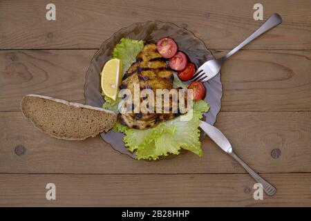 Bistecche di maiale grigliate su un piatto di lattuga, pomodori al limone e ciliegia con forchetta, coltello e pane nero casereccio a fette su tavole di legno sullo sfondo. H Foto Stock