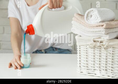 Primo piano di mani femminili versando detersivo liquido per bucato in tappo su tavola bianca rustica con asciugamani in cesto di vimini Foto Stock