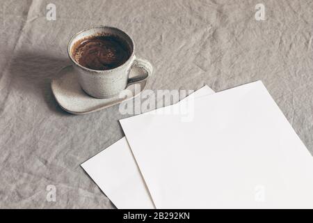 Tazza di caffè e fogli di carta vuoti. Moody colazione tavolo cancelleria mockup scena. Tovaglia di lino beige sullo sfondo. Sparse ancora la vita Foto Stock