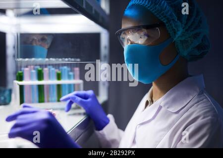 Ritratto della vista laterale dello scienziato femminile che indossa maschera facciale e equipaggiamento protettivo lavorando alla ricerca in laboratorio, copia spazio Foto Stock