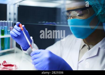 Ritratto della vista laterale di una donna scienziato che indossa maschera e un dispositivo di protezione che fa cadere i campioni di sangue nelle provette mentre si lavora alla ricerca in laboratorio, Foto Stock