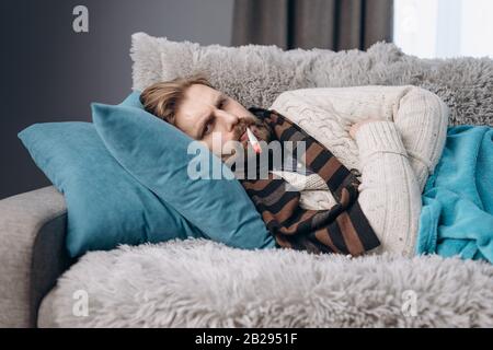 Malata in abiti caldi a casa sdraiata sul divano del soggiorno con termometro sulla bocca per misurare la temperatura corporea. Persona matura in maglia s Foto Stock