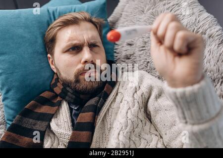 Vista dall'alto dell'uomo maturo in abiti caldi sentirsi deluso a causa della temperatura corporea che mostra il termometro. Uomo con bearded in sciarpa lavorato a maglia relaxi Foto Stock