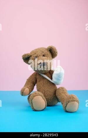 buffo orsacchiotto marrone vintage con zampa riavvolta con bendaggio di garza bianca seduta su sfondo blu, concetto di lesioni nei bambini o negli animali Foto Stock