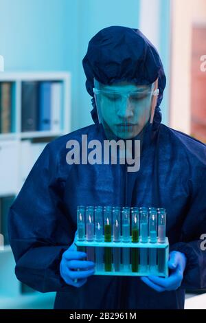 Ritratto verticale dello scienziato in costume protettivo completo che tiene provette di prova mentre si lavora alla ricerca in laboratorio Foto Stock