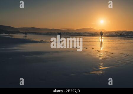 Silhouette di surfista persone che portano la loro tavola da surf sulla spiaggia al tramonto sulla riva del mare vicino a Bilbao in Spagna. Filtro vintage Foto Stock