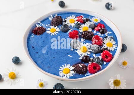 Ciotola frullato o Nizza crema fatta di blu spirulina, frutti di bosco surgelati, banana e noce di cocco con i fiori di camomilla su marmo bianco tavola. una sana prima colazione Foto Stock