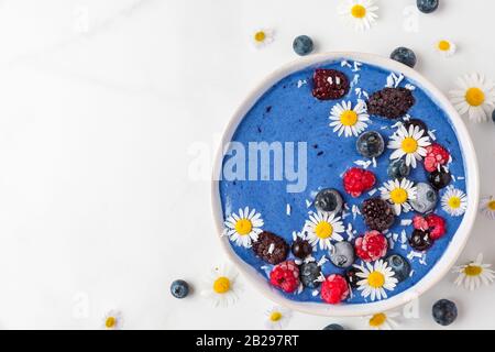 Ciotola frullato o Nizza crema fatta di blu spirulina, frutti di bosco surgelati, banana e noce di cocco con i fiori di camomilla su marmo bianco tavola. una sana prima colazione Foto Stock