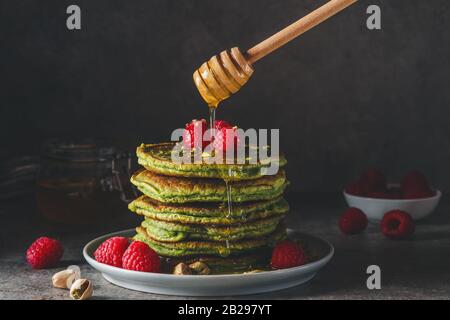 Frittelle Matcha verdi con lamponi freschi, pistacchi e miele che scorre su sfondo scuro. Chiave bassa. Colazione sana dessert. Tonalità vintage Foto Stock