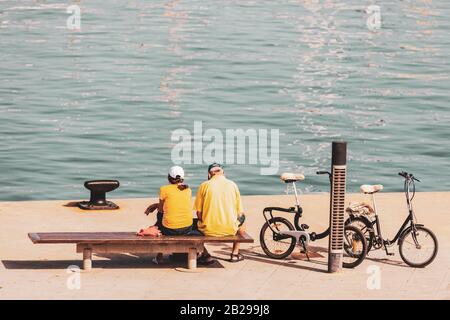 Donna anziana e uomo che indossano magliette e berretto gialli, seduti su una panchina del porto accanto alle loro biciclette pieghevoli Foto Stock