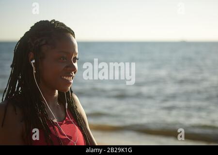 Ritratto di allegra giovane donna Nera ascoltando musica in cuffia e godendo della bellezza delle onde marine Foto Stock