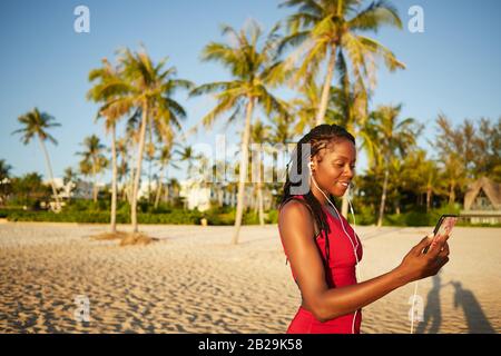 Donna sorridente in forma guardare video di allenamento sullo smartphone prima di iniziare ad allenarsi sulla spiaggia di sabbia Foto Stock