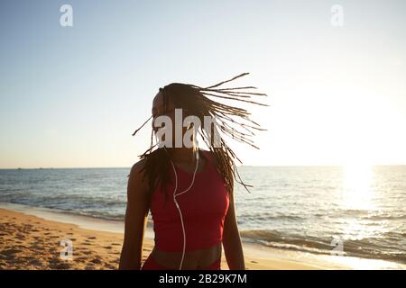 Felice sorridente giovane donna nera che balla e salta sulla spiaggia alla musica nelle sue cuffie Foto Stock