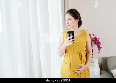 Giovane vietnamita incinta bella donna che tiene smartphone e tocca il suo ventre e guardando attraverso la finestra Foto Stock