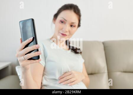 Bella giovane donna che tocca la pancia quando si prende selfie a casa, concentrarsi sullo smartphone Foto Stock