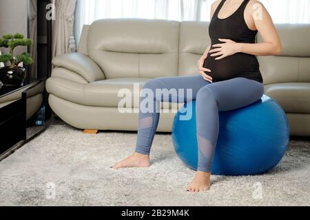 Immagine ritagliata di una donna in gravidanza sottile che tocca il ventre quando si riposa sulla palla yoga dopo aver lavorato fuori Foto Stock