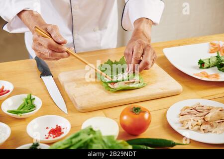 Mani di uomo che fa rotoli di lattuga con pollo, formaggio grattugiato e peperone Foto Stock