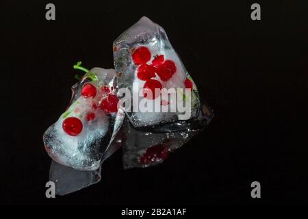 Bacche di ribes rosso congelate in blocchi di ghiaccio con riflessione su sfondo scuro. Ghiaccio incrinato. Primo piano, messa a fuoco selettiva Foto Stock