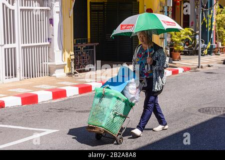 Una donna spinge un carretto a mano lungo un treet mentre tiene un ombrello, la città vecchia di Phuket, Thailandia Foto Stock