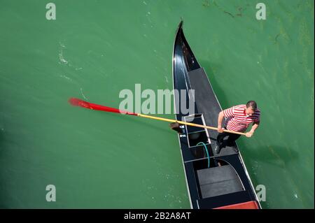 Venezia - 24 APRILE 2013: Gondoliere con camicia tradizionale a righe rosse che ranghi la sua gondola lungo un canale in un uniforme regolamentare di pantaloni neri. Foto Stock