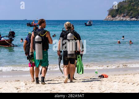 Quattro subacquei con vasche subacquee camminano verso il mare a Kata Beach, Phuket, Thailandia Foto Stock