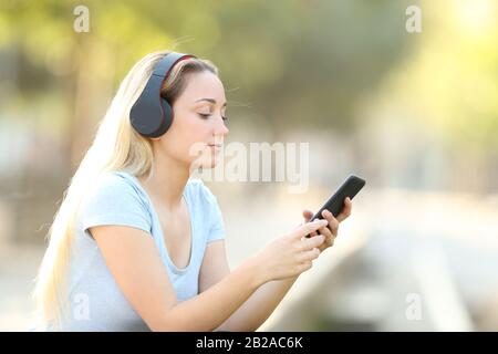 Ragazza giovane bionda che ascolta la musica sul suo telefono con le cuffie in un parco Foto Stock