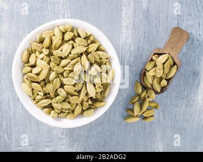 Cardamomo verde speziato (Elettraria cardamomum) in una ciotola bianca in ceramica con paletta in legno su fondo grigio in cemento. Il concetto di nutrizione adeguato Foto Stock