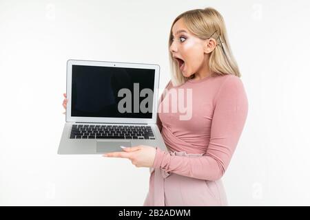 sorpresa ragazza bionda in vestiti rosa tiene un portatile con lo schermo in avanti con un vuoto vuoto su uno sfondo bianco studio Foto Stock