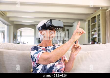 Vista frontale di una donna caucasica senior in un visore VR, toccando uno schermo virtuale con il dito seduto su un divano nel soggiorno, sorridendo. Famiglia en Foto Stock