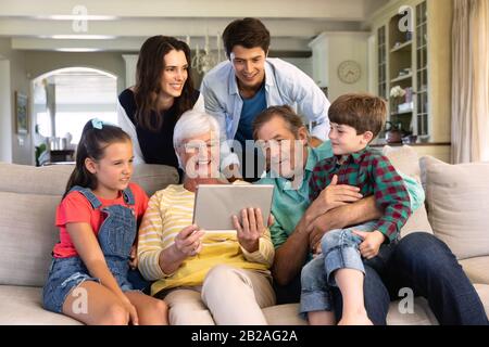 Vista frontale di sei membri di una famiglia caucasica multi-generazione a casa su un divano nel soggiorno, riuniti intorno alla nonna che sta tenendo Foto Stock