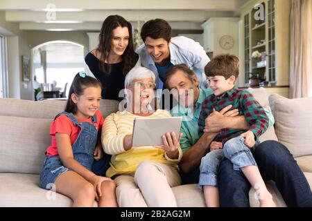 Vista frontale di sei membri di una famiglia caucasica multi-generazione a casa su un divano nel soggiorno, riuniti intorno alla nonna che sta mostrando Foto Stock