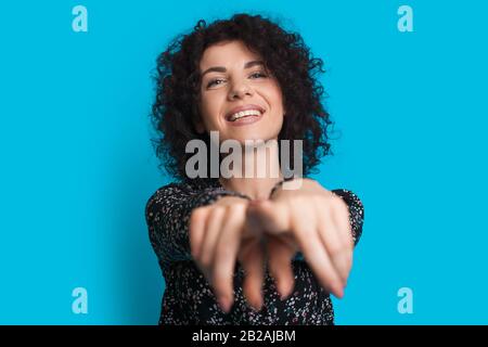 Curly capelli caucasici ragazza sta puntando alla macchina fotografica mentre sorridendo felicemente su una parete blu Foto Stock