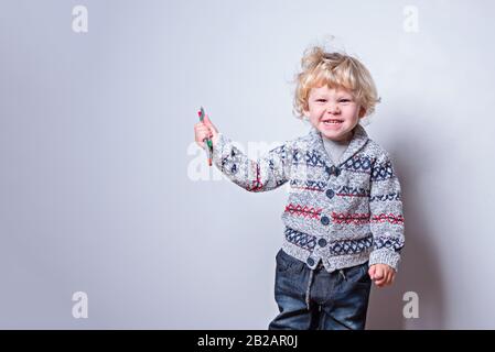 Un ragazzino con capelli ricci biondi tiene in mano matite multicolore e sorridente con i denti. Su sfondo bianco. Spazio di copia. Inserire il testo. Foto Stock