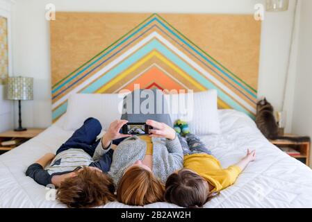Madre sdraiata a letto con due bambini che prendono un selfie Foto Stock
