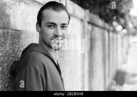 Felice giovane uomo barbuto sorridere mentre indossa felpa con cappuccio rosso e poggiante sul vecchio muro di cemento nelle strade all'aperto Foto Stock