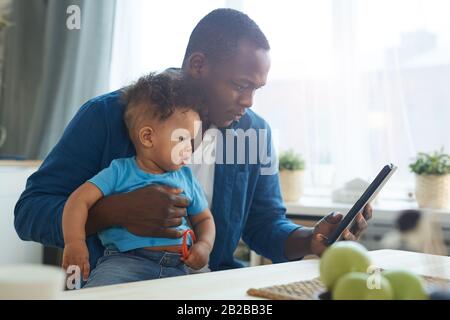 Ritratto della vista laterale di uomo afro-americano che tiene il bambino mentre usando il tablet digitale nell'interno domestico, copia spazio Foto Stock