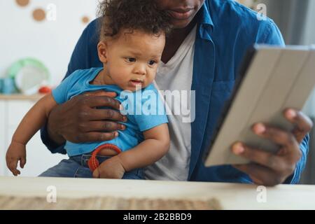 Ritratto corto di uomo afro-americano che tiene il bambino mentre usando il tablet digitale nell'interno domestico, copia lo spazio Foto Stock