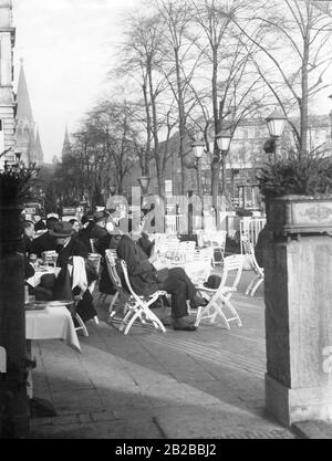 Gli ospiti possono sedersi in una caffetteria di strada a Berlino sul Kurfuerstendamm. Viene fotografata attraverso due colonne con canali di piante su di esse. Indossa i ghette sopra le scarpe. Sullo sfondo si può vedere la Chiesa commemorativa dell'Imperatore Guglielmo. Foto Stock