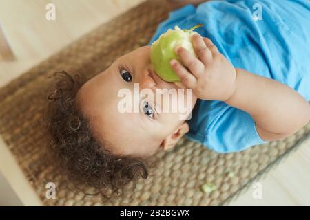 Sopra il ritratto di vista della razza cute mixed toddler che mangia la mela verde mentre osserva la macchina fotografica, lo spazio della copia Foto Stock