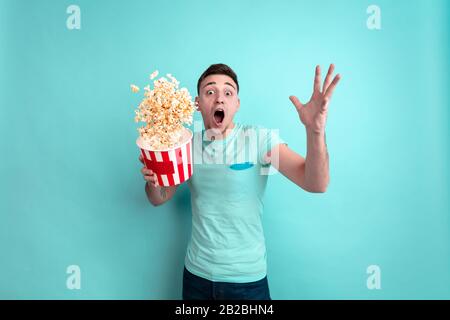 Pazzo scioccato, battenti popcorn. Ritratto del giovane caucasico isolato su sfondo blu studio. Bel modello maschile in stile casual. Concetto di emozioni umane, espressione facciale, vendita, pubblicità, cinema. Foto Stock