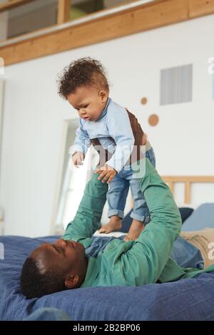 Ritratto verticale di padre afro-americano felice che gioca con il figlio del bambino mentre posa sul letto a casa Foto Stock