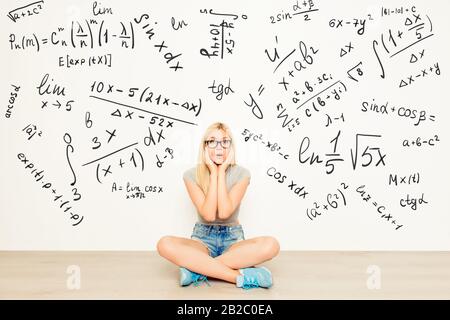 attraente ragazza scioccata in occhiali seduti a terra vicino a parete con formule matematiche, gambe incrociate e viso toccante Foto Stock