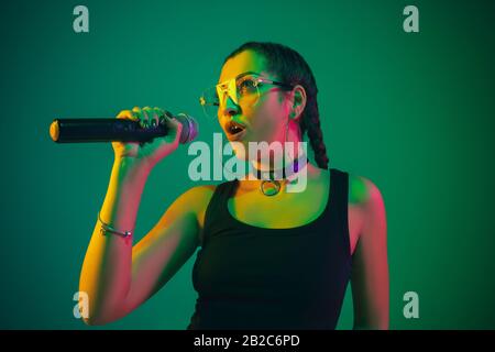 Ritratto femminile caucasico isolato su sfondo verde studio in luce neon. Bella femmina modello in nero con microfono. Concetto di emozioni umane, espressione facciale, annuncio, musica, arte Foto Stock