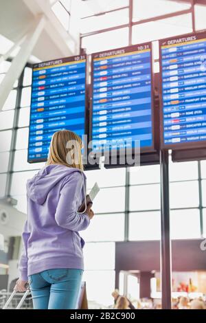 Vista posteriore della giovane adolescente che tiene il passaporto e la carta d'imbarco mentre guarda il suo volo sul monitor Foto Stock